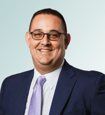 George Chause Jeffrey Ebel, dressed in a dark blue suit, white shirt, and light purple tie, is smiling warmly. He has short dark hair and is wearing glasses. The background features a light gradient blue that complements his professional appearance.
