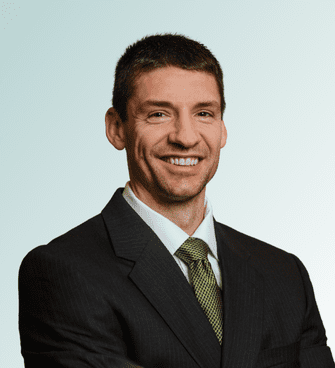 Jeffrey Ebel Jeffrey Ebel, a man with short dark hair and a light beard, is smiling in a dark suit, white shirt, and green tie. He stands confidently against a light gradient background, facing the camera with his arms crossed.