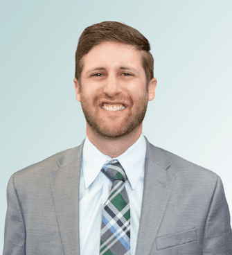 Jordan A man with short brown hair and a beard, likely Dr. Jordan Feierman, is smiling while wearing a light gray suit, a light blue dress shirt, and a plaid tie with shades of green, blue, and white. The background transitions from light blue to white.