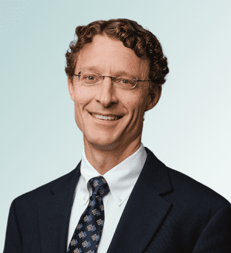 Nathan Hunt A man with curly hair and glasses, possibly Dr. Jeffrey Ebel, is wearing a dark suit, white shirt, and patterned tie. He is smiling and standing against a light blue gradient background.
