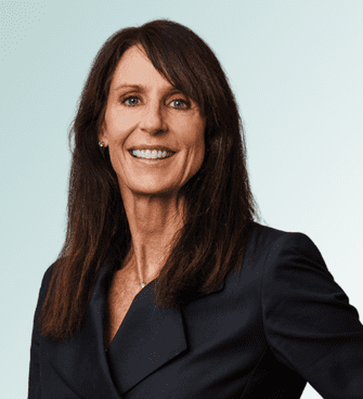 Niki Vischer Johnson A woman with long brown hair and a friendly smile, Dr. Niki Vischer, MD, is wearing a dark blazer. She stands against a light, gradient background.