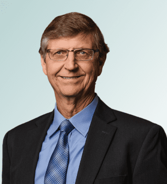 A man with short, light brown hair and glasses is wearing a blue dress shirt, a patterned blue tie, and a dark suit jacket. He is smiling against a plain, light blue background. Meet Ray van den Hoven, MD.
