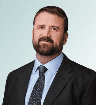 Dr. Riley Hale, a man with short brown hair and a beard, is wearing a dark suit, light blue dress shirt, and patterned tie. He is posing against a light blue gradient background.