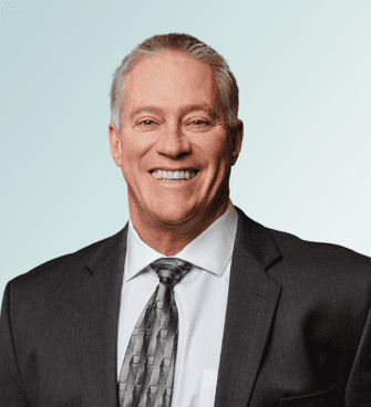 A man with short gray hair and a broad smile, identified as Jeffrey Ebel, is wearing a dark suit, white dress shirt, and patterned tie. He stands against a light blue gradient background.
