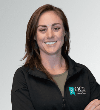 Copy of OCR_Trauma Team (1) A woman with shoulder-length brown hair is smiling and standing with her arms crossed. She is wearing a black jacket with the text “OCR” and a ribbon graphic on the left side. The background is a plain light gray color. Alexandria Stedry, PA-C, exudes confidence and professionalism in her attire.