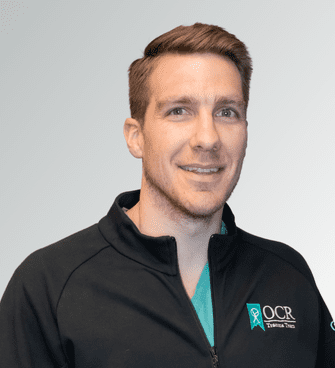 Copy of OCR_Trauma Team A man with short brown hair smiles at the camera. He is wearing a black jacket with the acronym "OCR" and the phrase "Orthopaedic & Spine" on it, along with a green shirt underneath. The background is a gradient of light gray. This is Isaac DuCharme, PA-C.
