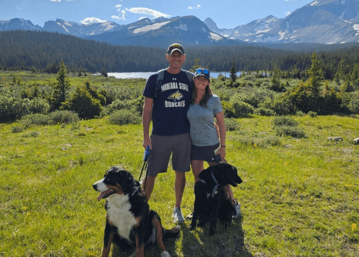 a man and a woman standing on green grass in front of a mountain with two dogs a man and a woman standing on green grass in front of a mountain with two dogs