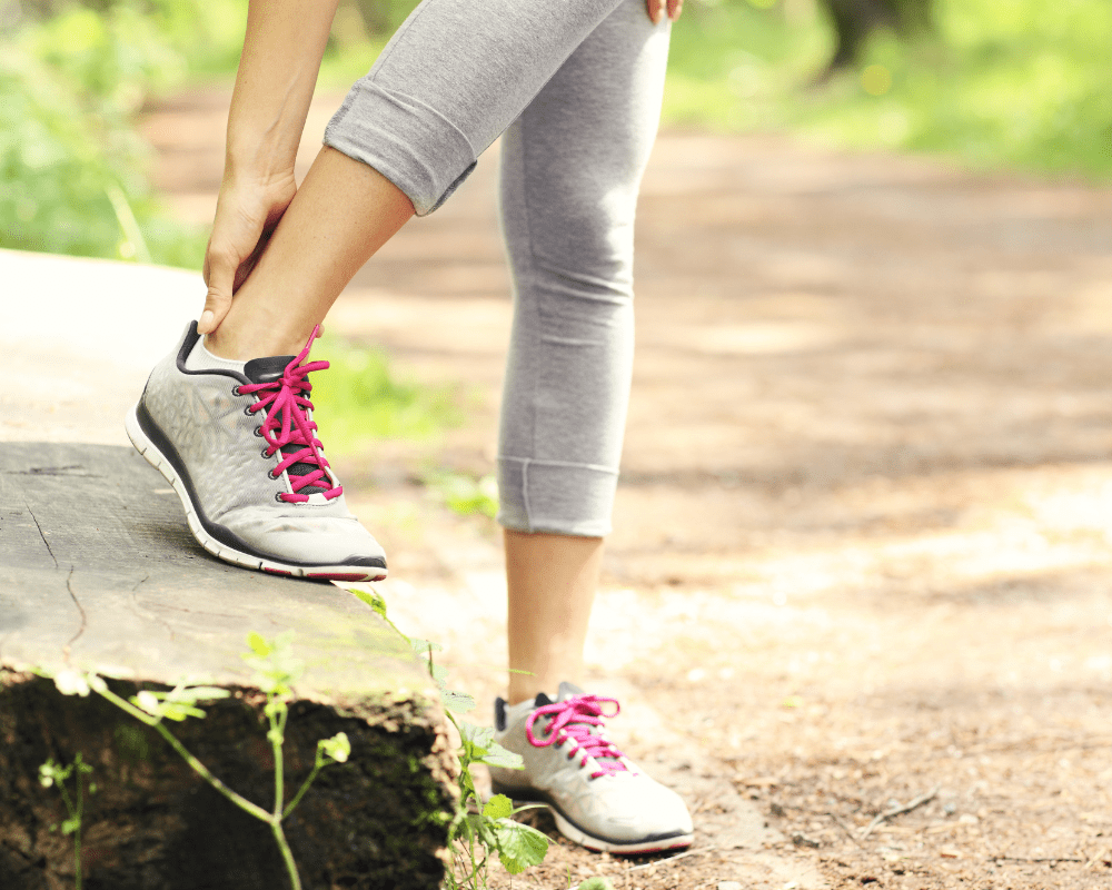 A person in gray athletic leggings and sneakers with pink laces, using one leg to rest on a tree stump while stretching their calf muscle on a wooded trail. The background shows a dirt path lined with greenery and dappled sunlight. A person in gray athletic leggings and sneakers with pink laces, using one leg to rest on a tree stump while stretching their calf muscle on a wooded trail. The background shows a dirt path lined with greenery and dappled sunlight.