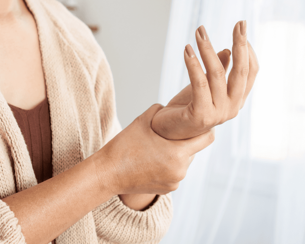 A person wearing a beige cardigan and brown shirt holds their right wrist with their left hand, appearing to examine or massage it. The background is softly lit with light-colored curtains. A person wearing a beige cardigan and brown shirt holds their right wrist with their left hand, appearing to examine or massage it. The background is softly lit with light-colored curtains.