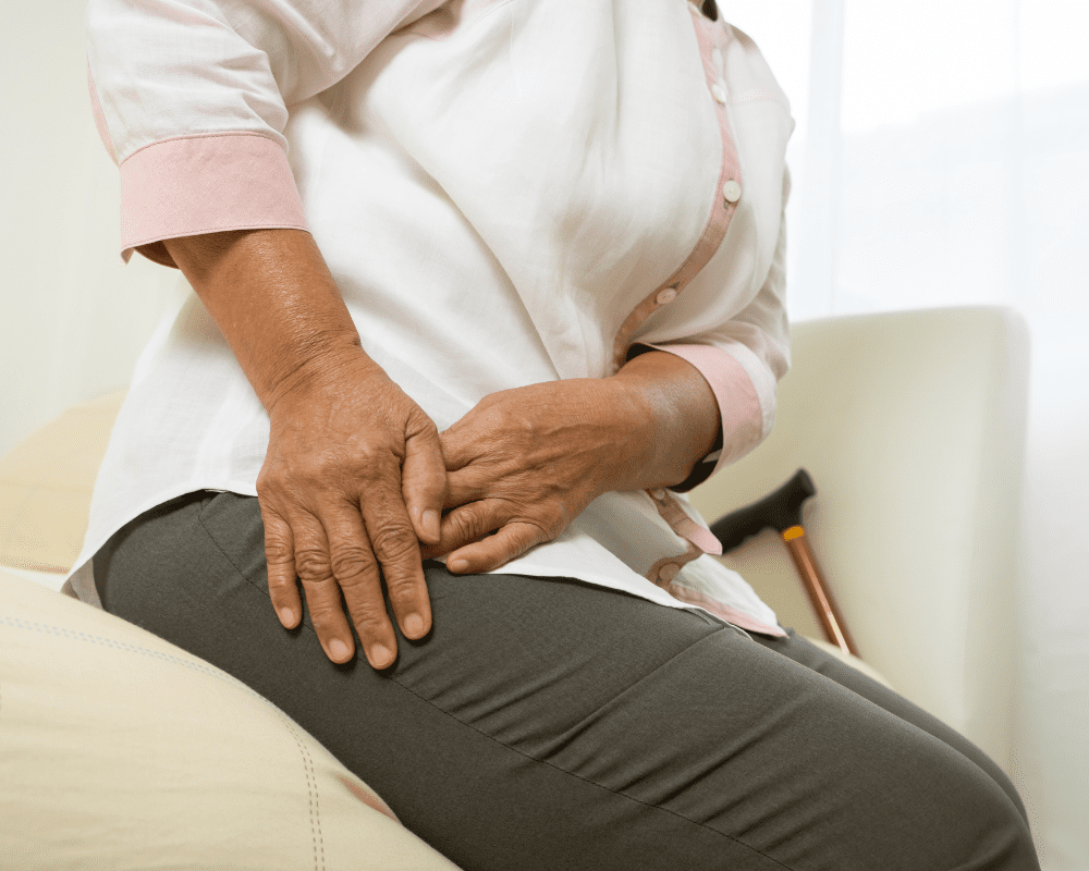A person wearing a white and pink shirt and gray pants sits on a cushion while clutching their right hip with both hands, suggesting discomfort or pain. A cane rests beside them. A person wearing a white and pink shirt and gray pants sits on a cushion while clutching their right hip with both hands, suggesting discomfort or pain. A cane rests beside them.