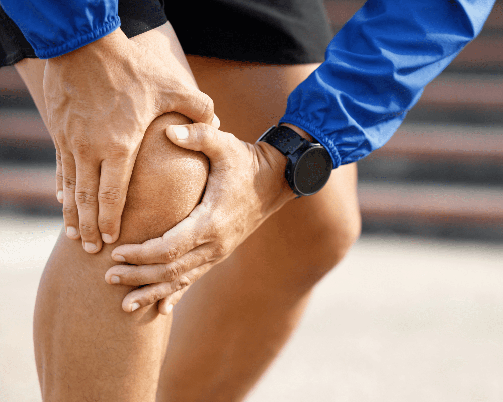 A person wearing a blue long-sleeve shirt and black shorts is holding their right knee with both hands, indicating potential pain or injury. They are wearing a black watch on their left wrist. The background is blurred, suggesting an outdoor setting. A person wearing a blue long-sleeve shirt and black shorts is holding their right knee with both hands, indicating potential pain or injury. They are wearing a black watch on their left wrist. The background is blurred, suggesting an outdoor setting.