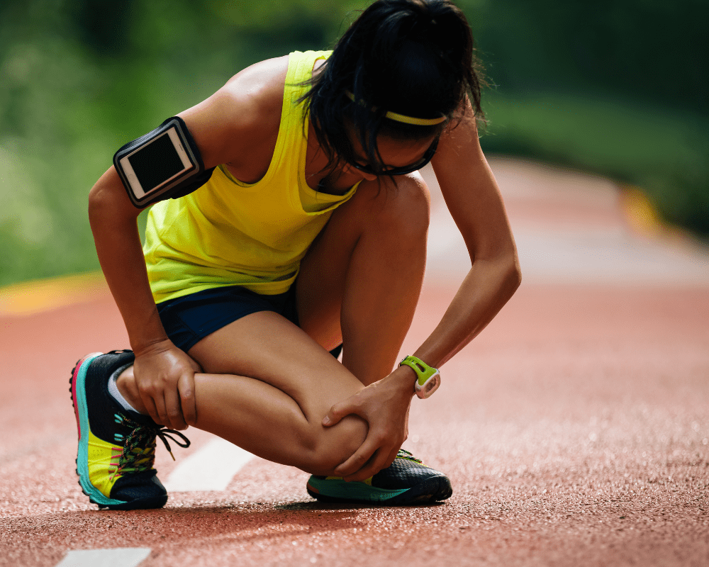 A person dressed in athletic gear crouches down on a running track, holding their lower leg with a pained expression. They are wearing a yellow tank top, dark shorts, colorful running shoes, a fitness tracker, and an armband for a device. A person dressed in athletic gear crouches down on a running track, holding their lower leg with a pained expression. They are wearing a yellow tank top, dark shorts, colorful running shoes, a fitness tracker, and an armband for a device.