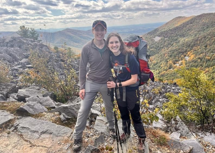 Dr. Thorne Hiking Two hikers stand on a rocky trail with scenic mountains in the background. Both are smiling and wearing hiking gear; one has a backpack and trekking poles. The landscape features autumn-colored foliage under a partly cloudy sky.