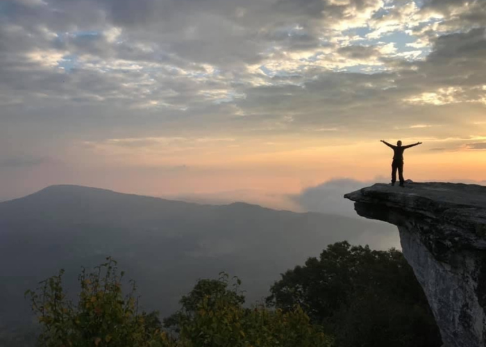 Sunset A person stands with arms outstretched on the edge of a rocky cliff overlooking a vast, mountainous landscape during sunrise or sunset. The sky is filled with clouds, casting a serene light over the scene. Vegetation can be seen in the foreground.