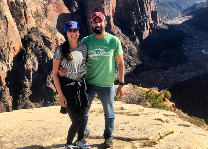 Dr. Suwak A couple stands on a rocky cliff with scenic canyon views in the background. The woman wears a blue cap and striped shirt, while the man wears a red cap and green t-shirt. They are both smiling and embracing each other amid the natural landscape.