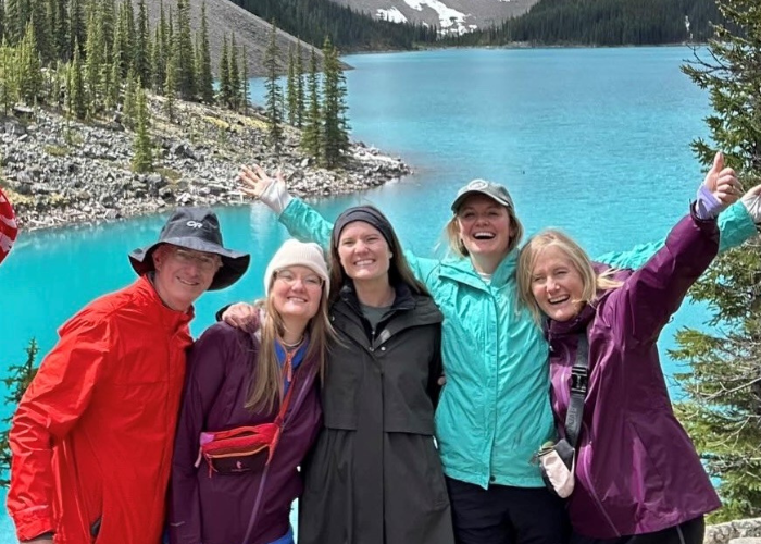 the FitzGibbons Family A group of five people, wearing colorful outdoor clothing, smile and pose cheerfully in front of a stunning turquoise lake surrounded by pine trees and mountains. The weather appears cool, and the group seems to be enjoying a nature adventure together.