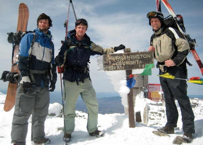 Dr. Alexander Mt. Washington Summit Three individuals dressed in winter gear stand at the snow-covered summit of a mountain next to a weathered sign that reads "Washington Summit" and other partially obscured text. Each person is holding skis or a snowboard. The sky is clear in the background.