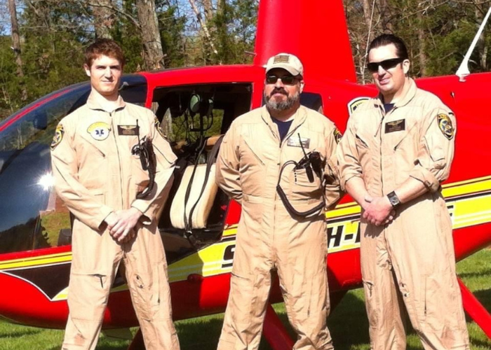 Dr. Alexander & Team Three men in tan flight suits stand in front of a red helicopter with "SOUTH" partially visible on the side. The men have various gear attached to their suits and wear sunglasses. The background features trees and a grassy area.