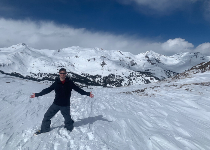Dr. Alexander in Snow A person wearing sunglasses, a black jacket, and gray snow pants stands with outstretched arms on a snow-covered landscape with mountains in the background under a partly cloudy sky.
