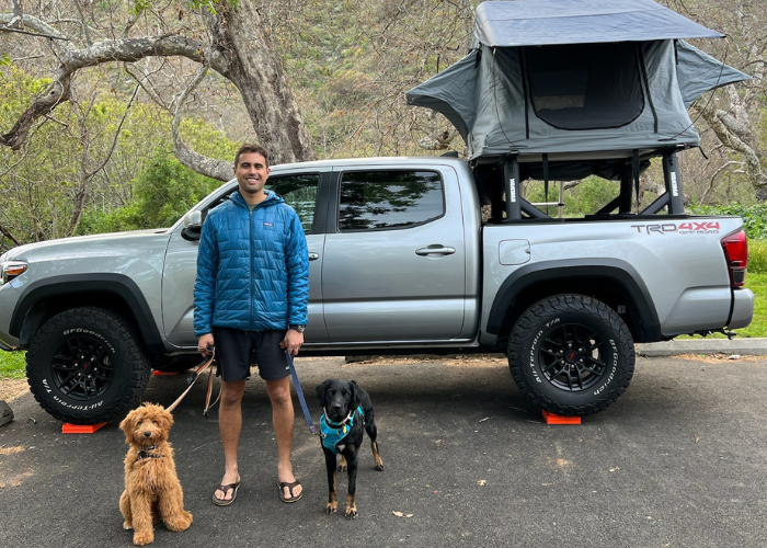 Dr. Spady & Dogs A man stands smiling next to a pickup truck with a rooftop tent. He holds leashes with two dogs: a fluffy brown dog on the left and a black dog with a blue bandana on the right. Trees and grassy areas are visible in the background.