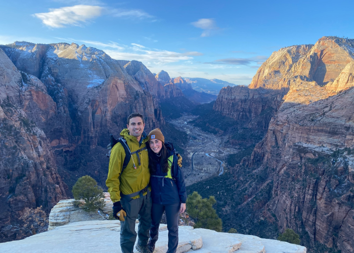 Dr. Spady Hiking A man and woman stand together on a rocky ledge with a vast canyon landscape in the background. They are both wearing hiking gear, including jackets and beanies. The sun is setting, casting a warm glow on the mountains and the valley below.