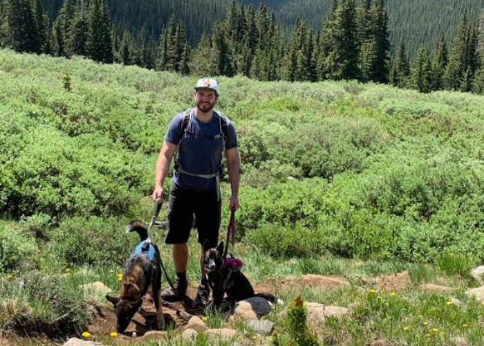 Dr. Torri & Dogs A person stands on a hiking trail in a lush, green mountainous area, holding the leashes of two dogs. The person wears a cap, a backpack, a blue t-shirt, and black shorts. One dog seems to be sniffing the ground, while the other is sitting attentively. Pine trees are visible in the background.