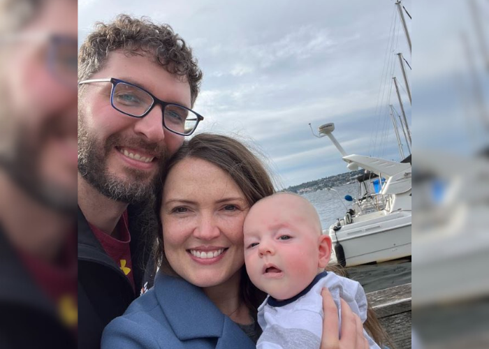 Dr. Torri & Family A man with glasses and a beard, a woman with long brown hair, and a baby are smiling in front of a marina with boats. The sky is cloudy. They appear to be enjoying a day out by the water.