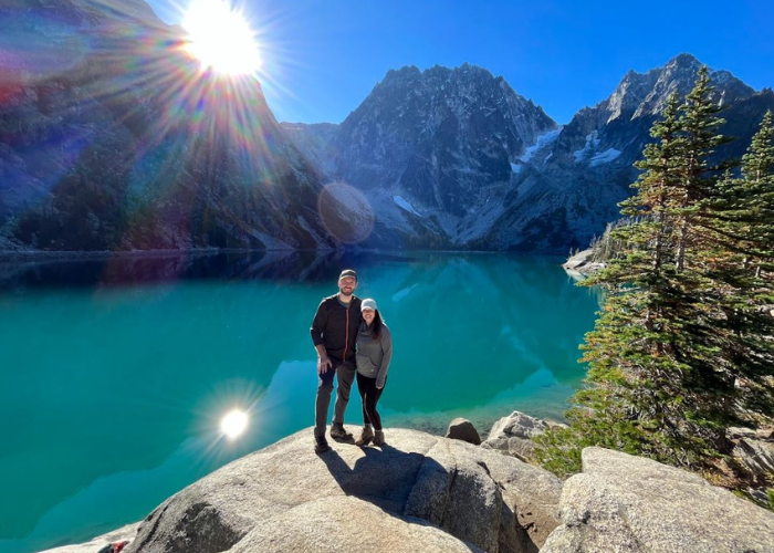 Dr. Torri Hiking A man and a woman stand in front of a crystal blue lake with mountains and trees behind them.