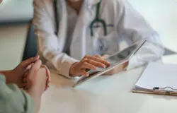 featured-appointments A doctor in a white coat uses a tablet during a consultation with a patient. The patient’s hands are clasped on the table, and a clipboard with papers is nearby.