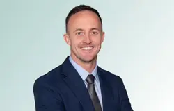 featured-mchugh Smiling man wearing a navy suit, light blue shirt, and gray tie, standing with arms crossed in front of a light gradient background.
