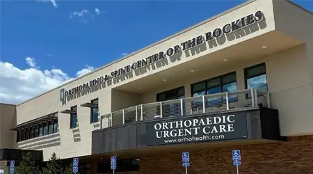 longmont-exterior A two-story medical building with signs reading "ORTHOPAEDIC & SPINE CENTER OF THE ROCKIES" and "ORTHOPAEDIC URGENT CARE" against a blue sky, with accessible parking spaces in front.