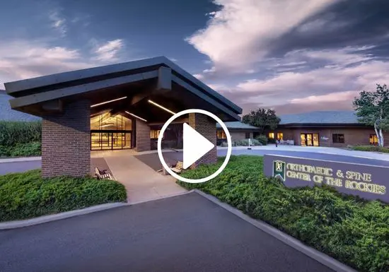ocr-video-home Exterior view of the Orthopaedic & Spine Center of the Rockies building at dusk, with warm lights glowing inside, landscaped grounds, and a prominent entrance under a large overhang. A play button icon is overlaid on the image.