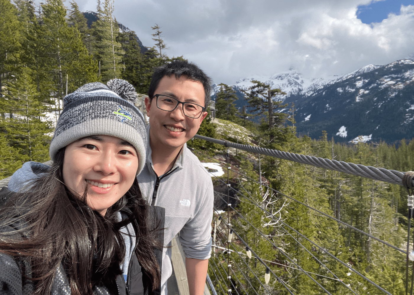 Ng 1 Two people in winter jackets and hats smile for a selfie on a suspension bridge, surrounded by pine trees and snow-capped mountains under a partly cloudy sky.