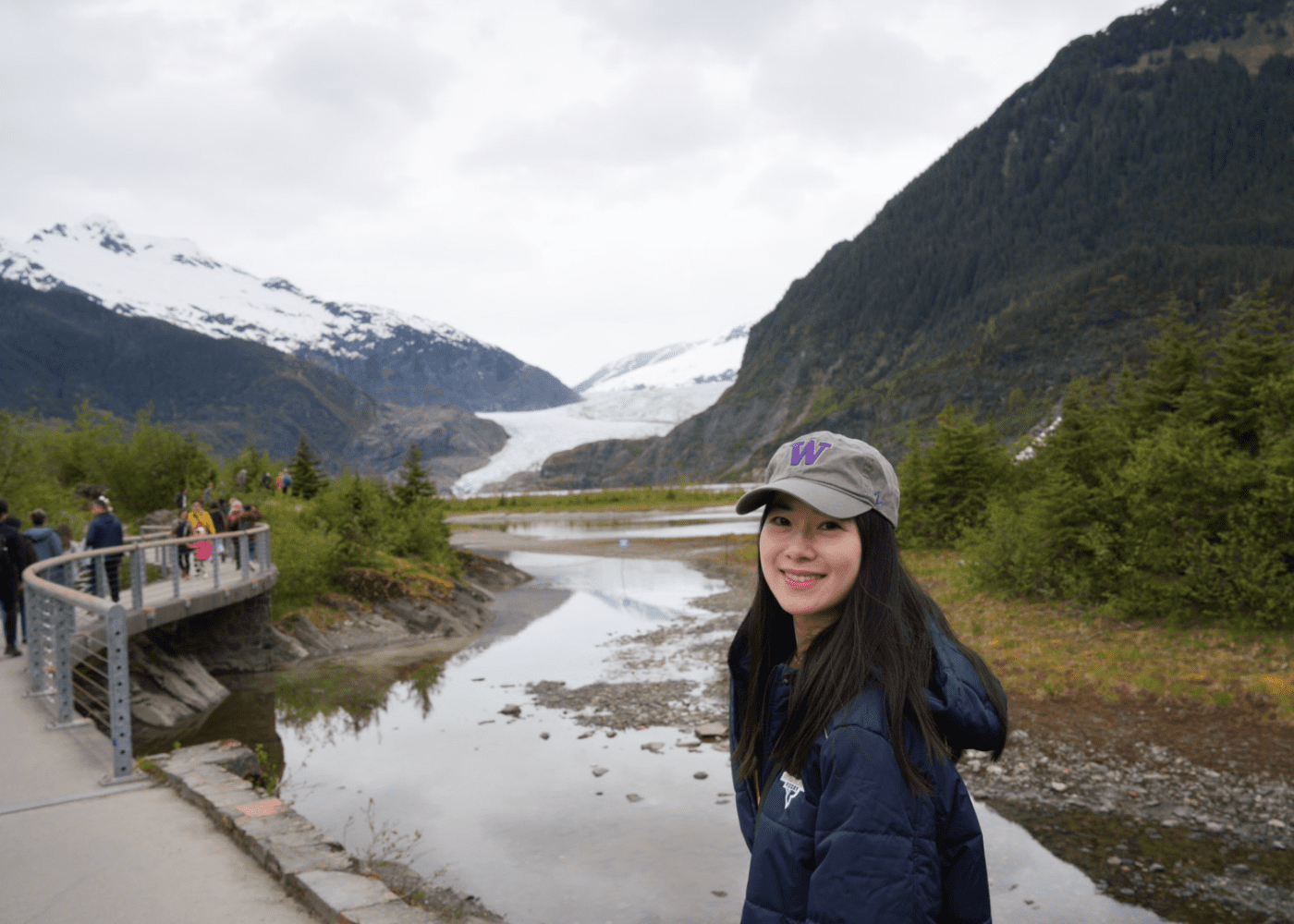 Ng 2 A woman in a navy jacket and cap smiles at the camera, standing near a mountain landscape with a river and glacier. People walk on a path to the left, and snow-capped mountains rise in the background.