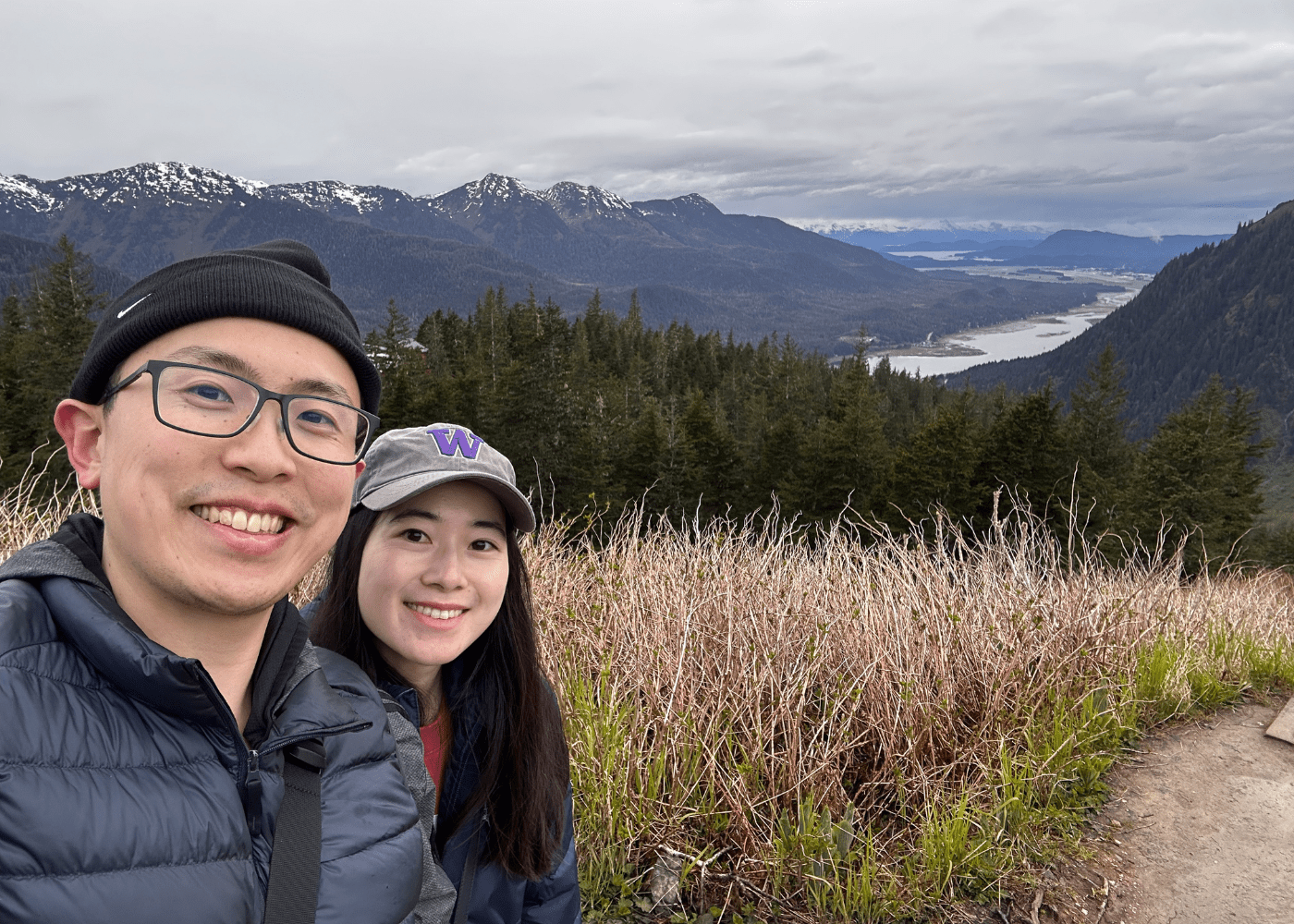 Ng 3 A man and a woman smile for a selfie on a hiking trail with snow-capped mountains, dense forest, and a river valley in the background under a cloudy sky.
