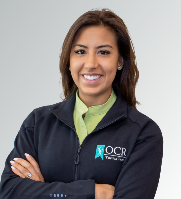 Gaby Londono (1) Gabriela Londono, PA-C, a woman with brown hair, smiles with arms crossed in a black "OCR Trauma Team" zip-up jacket and lime-green shirt, standing in front of a plain light gray background.