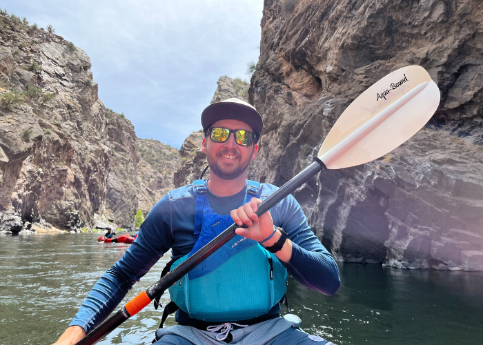Malesovas 1 A man wearing sunglasses, a cap, and a blue life vest smiles while kayaking through a rocky canyon on a sunny day, holding a paddle over the water.