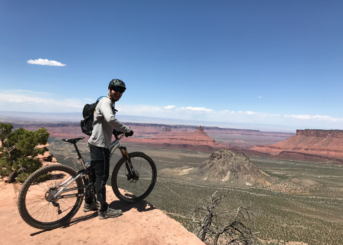 Malesovas 2 A person wearing a helmet and backpack stands beside a mountain bike on a rocky cliff, overlooking a vast desert landscape with mesas and buttes under a clear blue sky.