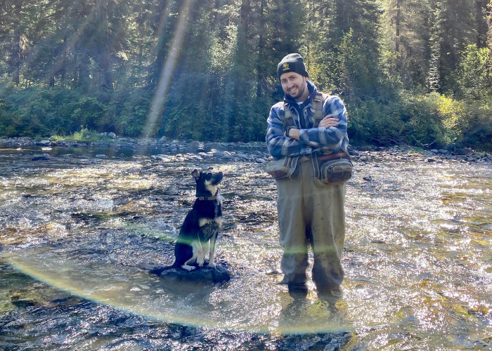 Malesovas 3 A man in outdoor gear stands in a shallow, sunlit river next to a black and white dog. Tall trees and greenery fill the background, with sunlight streaming through the forest.