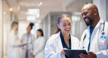 Two doctors in white coats smile and look at a tablet together in a bright hospital hallway, while two other pairs of medical professionals talk in the background.