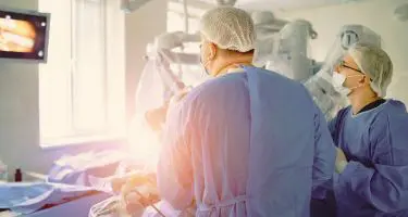 Two surgeons in blue scrubs, hair covers, and masks perform surgery in a brightly lit operating room, while viewing a monitor displaying a medical image.