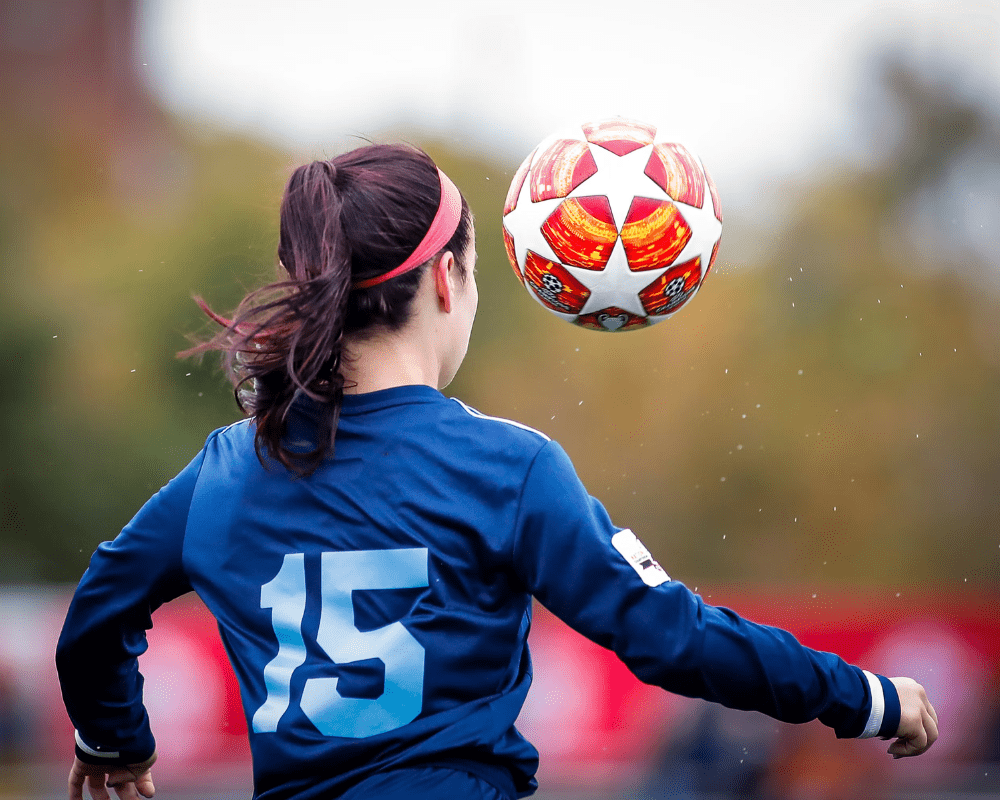 A soccer player wearing a blue jersey with the number 15 heads a soccer ball during a game. The player's back is facing the camera, and the background is blurred, focusing on the action.