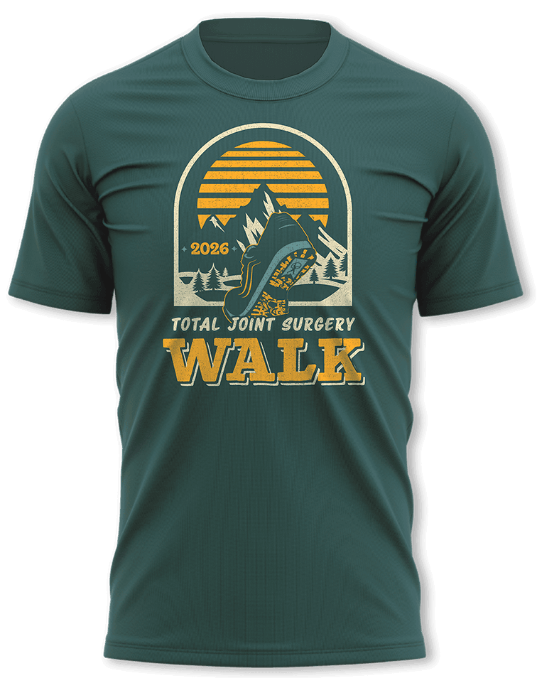 Green t-shirt with a graphic of a hiker, mountains, trees, and a sunset. Text reads “2026 Total Joint Surgery Walk.”.
