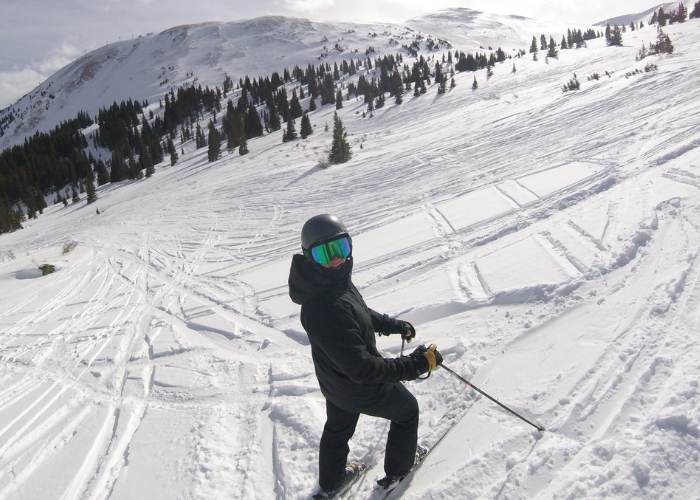 A skier wearing black gear and a helmet stands on a snowy mountain slope with ski tracks and trees in the background under a partly cloudy sky.