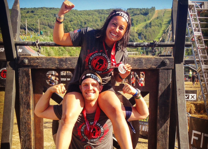Two people wearing medals and Spartan race shirts celebrate outdoors; the woman sits on the man’s shoulders, both smiling and flexing their arms. A race course and hills are visible in the background under a sunny sky.