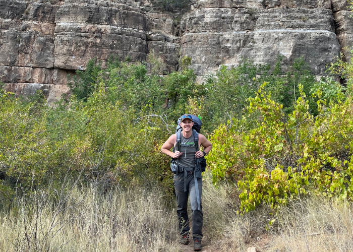 A person wearing a hat, sunglasses, and hiking gear walks on a trail surrounded by green bushes with a rocky cliff in the background.