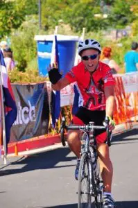 Steve in a red jersey and helmet waves while riding past a race barrier, celebrating his shoulder comeback. Other people and flags are visible in the background on a sunny day.