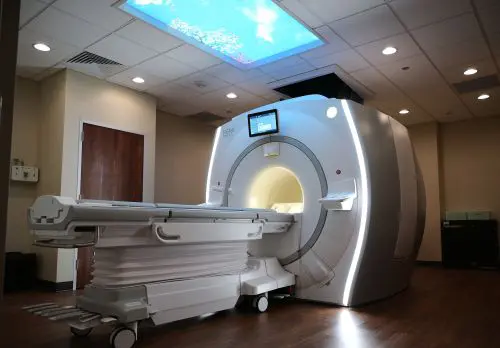 Longmont-MRI-web An MRI scanner with an extendable patient table in a medical examination room.