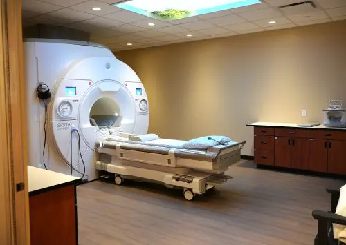 Loveland-MRI-web An MRI machine with a patient table in a medical examination room.
