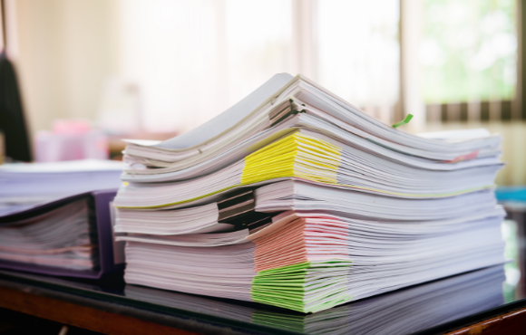 A stack of documents and files with colorful sticky notes and tabs sits on a desk in a well-lit office, indicating work or organization tasks.